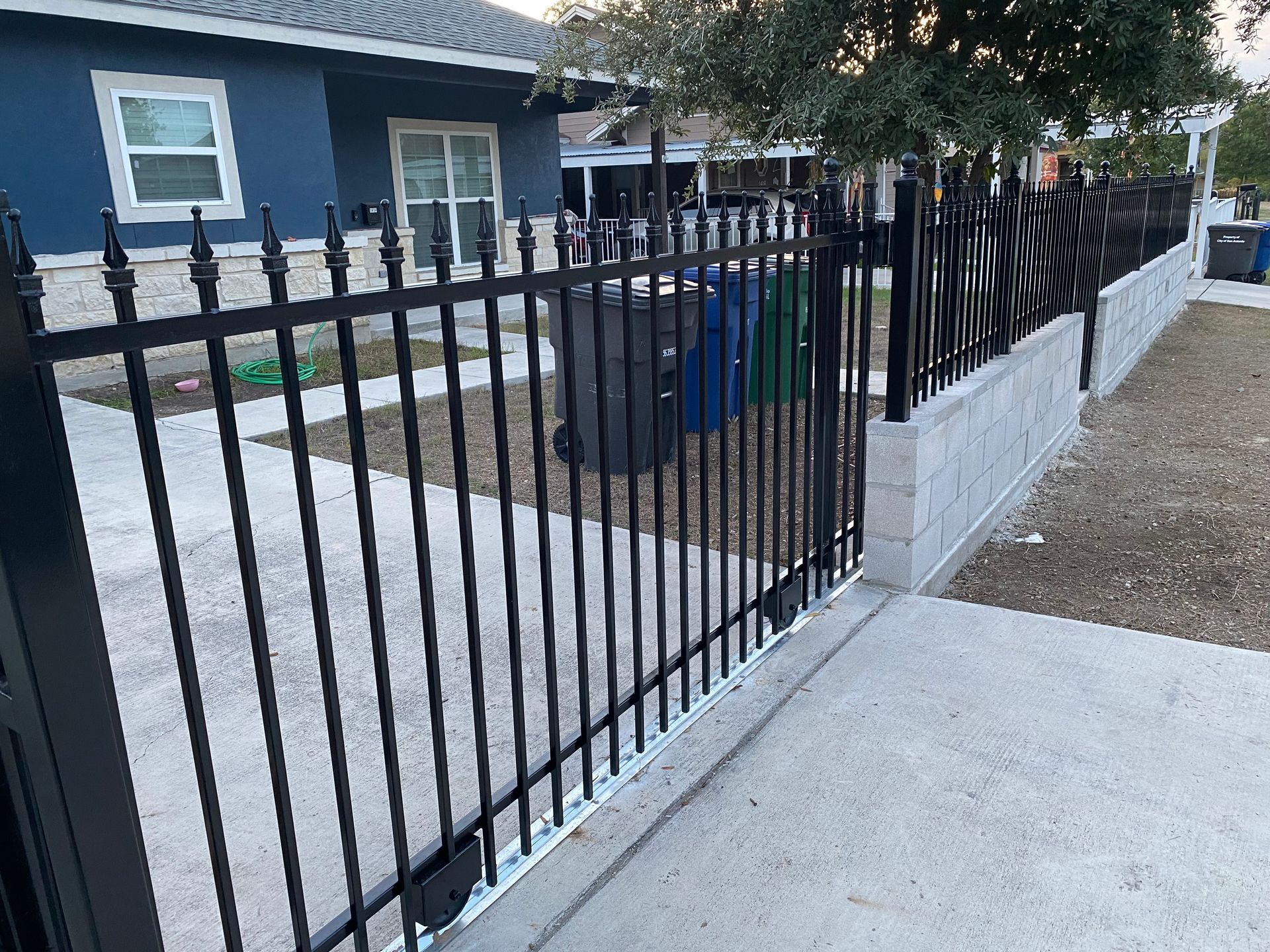 Black metal fence along a concrete sidewalk beside a blue house and driveway under trees