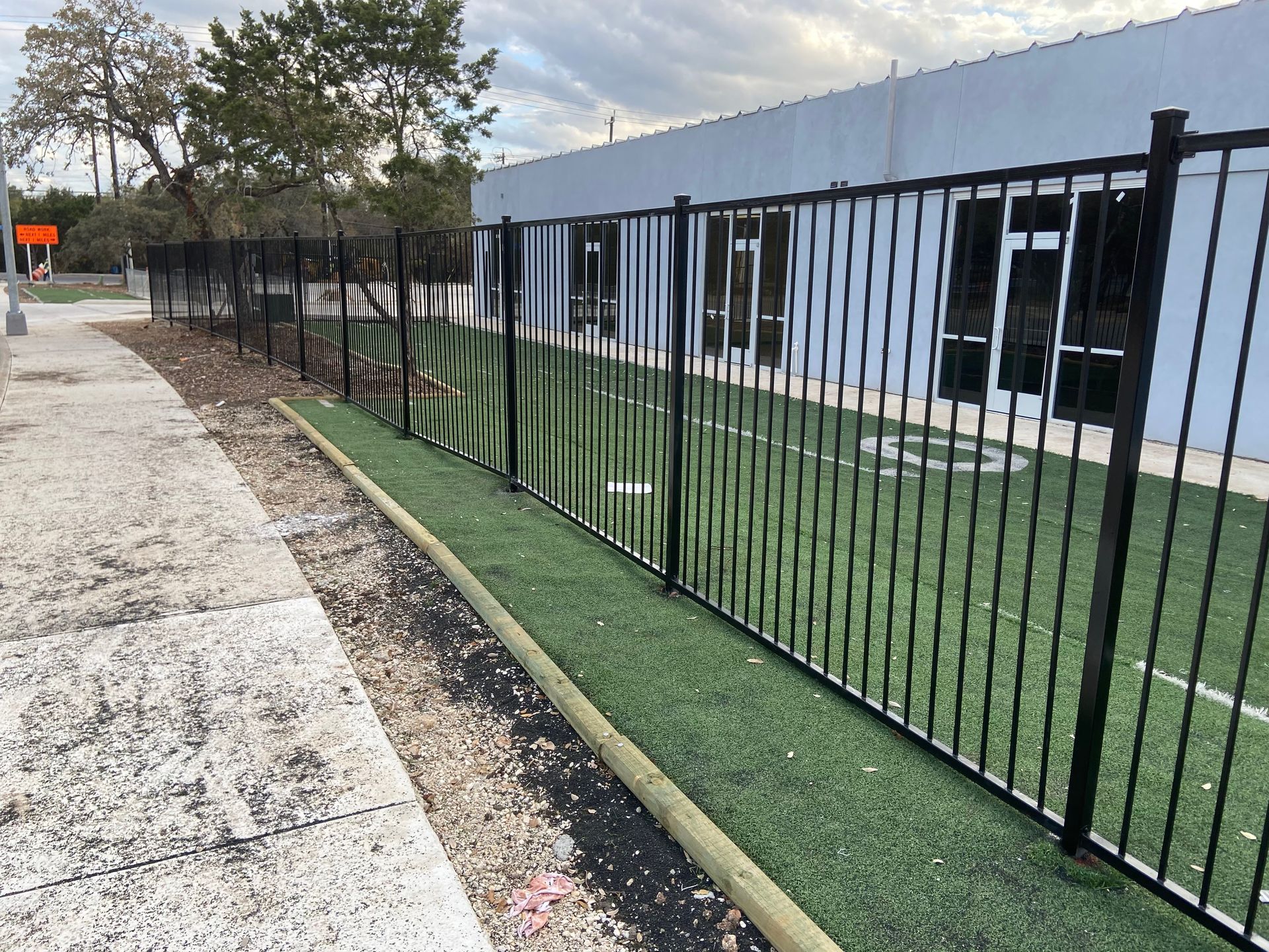 Black metal fence beside a building and walkway with artificial grass and trees along the side