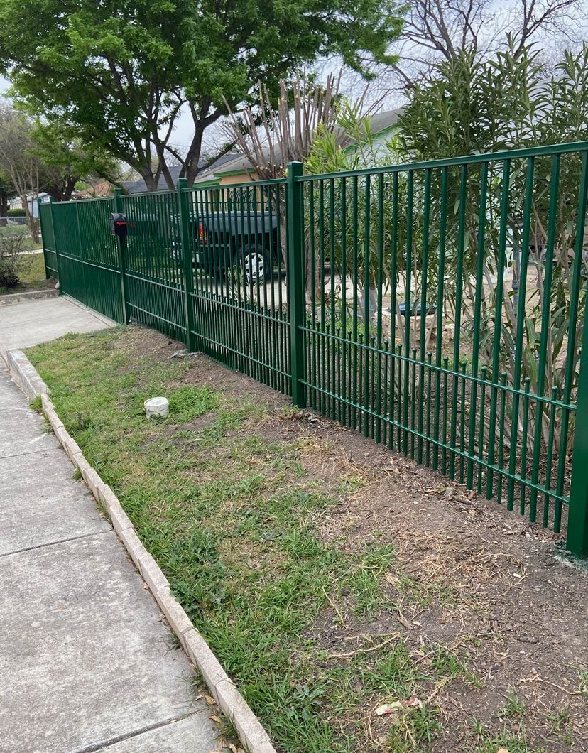Green metal fence beside a sidewalk and grassy strip with trees in a park-like area