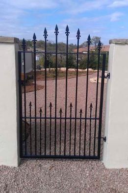Black wrought-iron gate between white walls, opening to a gravel driveway and landscaped yard.