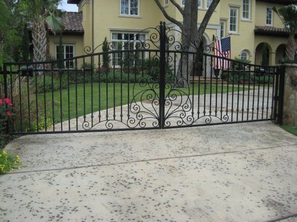 Black wrought-iron driveway gate in front of a yellow house with a green lawn