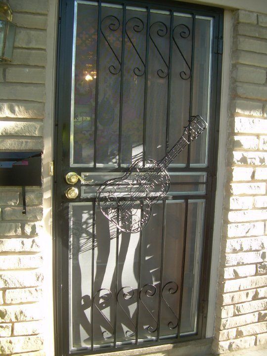 Black metal security door with decorative bars on a brick house exterior, lit by sunlight.