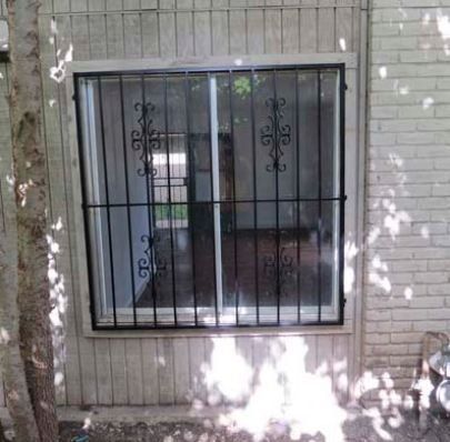 Window with black metal security bars set in a light brick wall, shaded by a tree outside.
