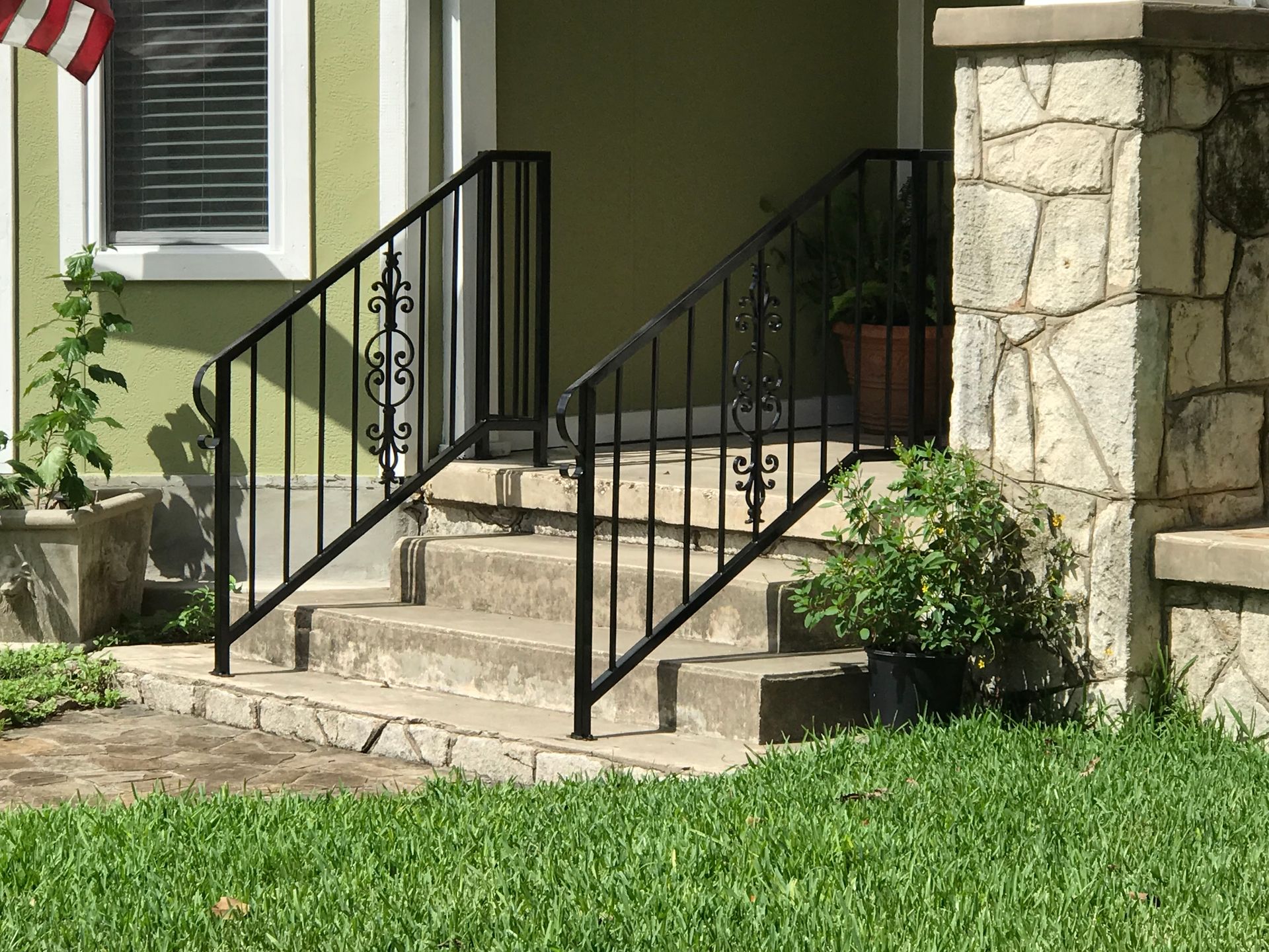 Front steps with black metal railings leading to a tan house, bordered by stone and green lawn