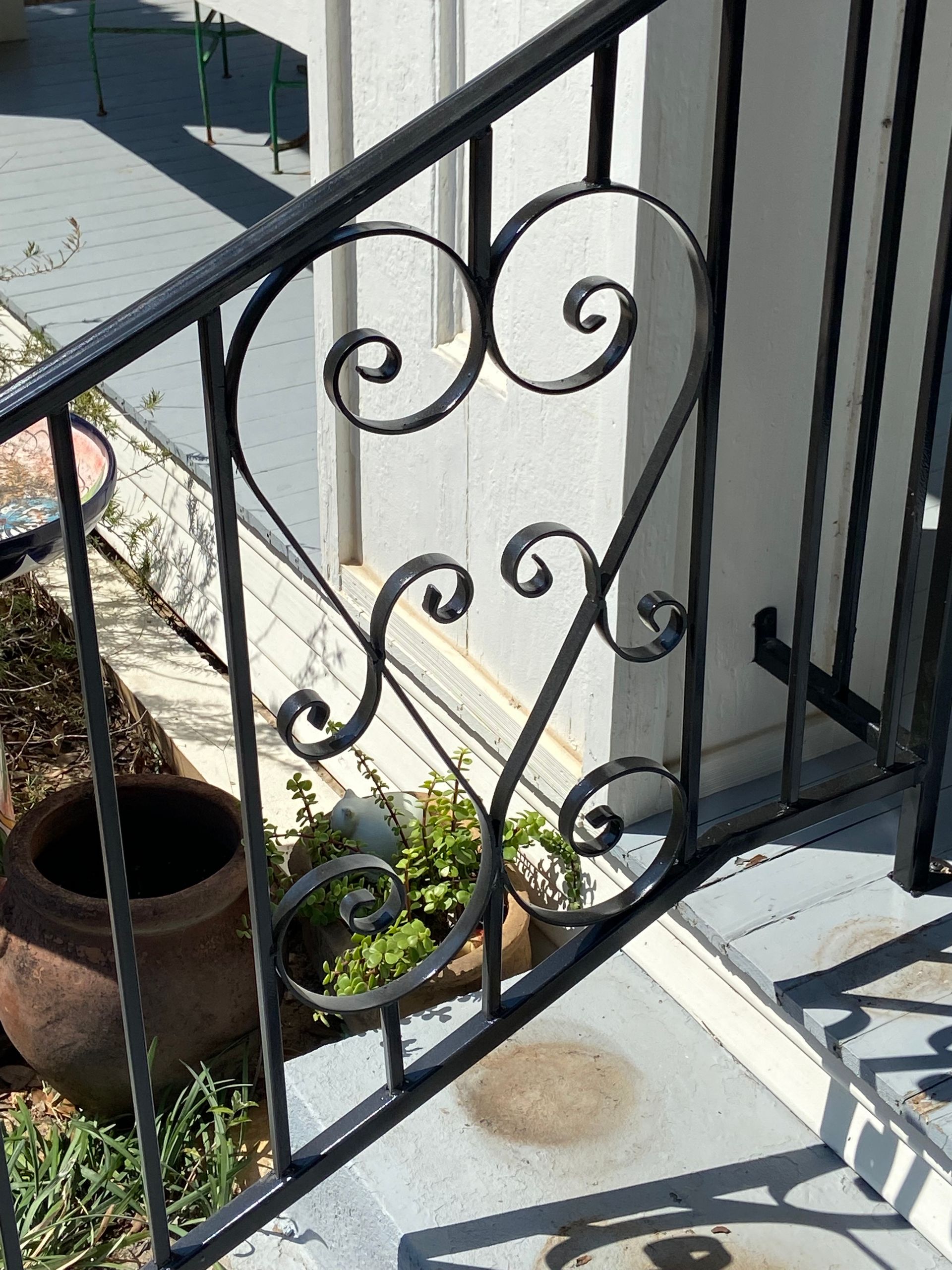 Black wrought-iron stair railing with decorative scrolls beside white steps and a small potted plant outdoors