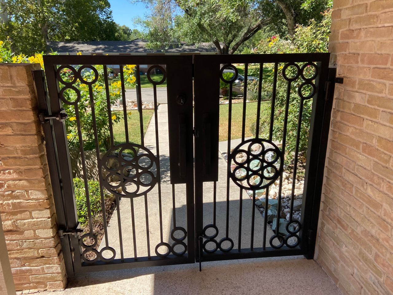 Black wrought-iron gate between brick walls beside a sunlit garden path