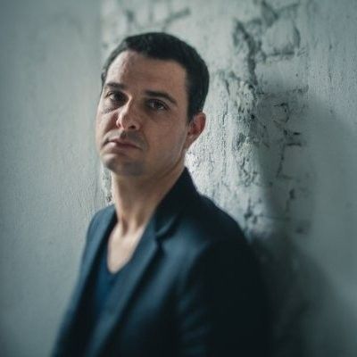 Um homem de terno está encostado em uma parede de tijolos.