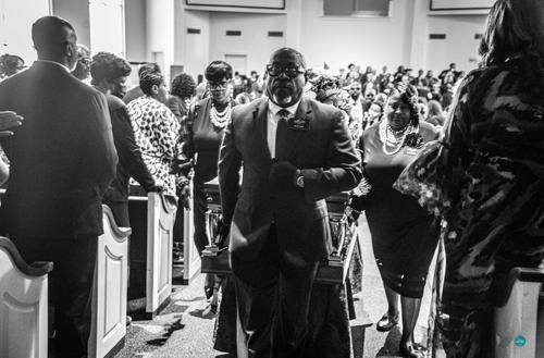 Black-and-white photo of a man in a suit walking down a church aisle during a crowded ceremony