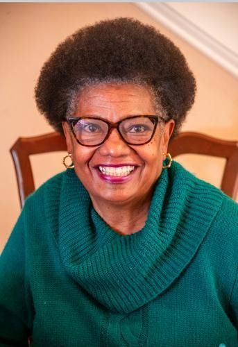 Smiling woman in green sweater and glasses seated indoors on a wooden chair