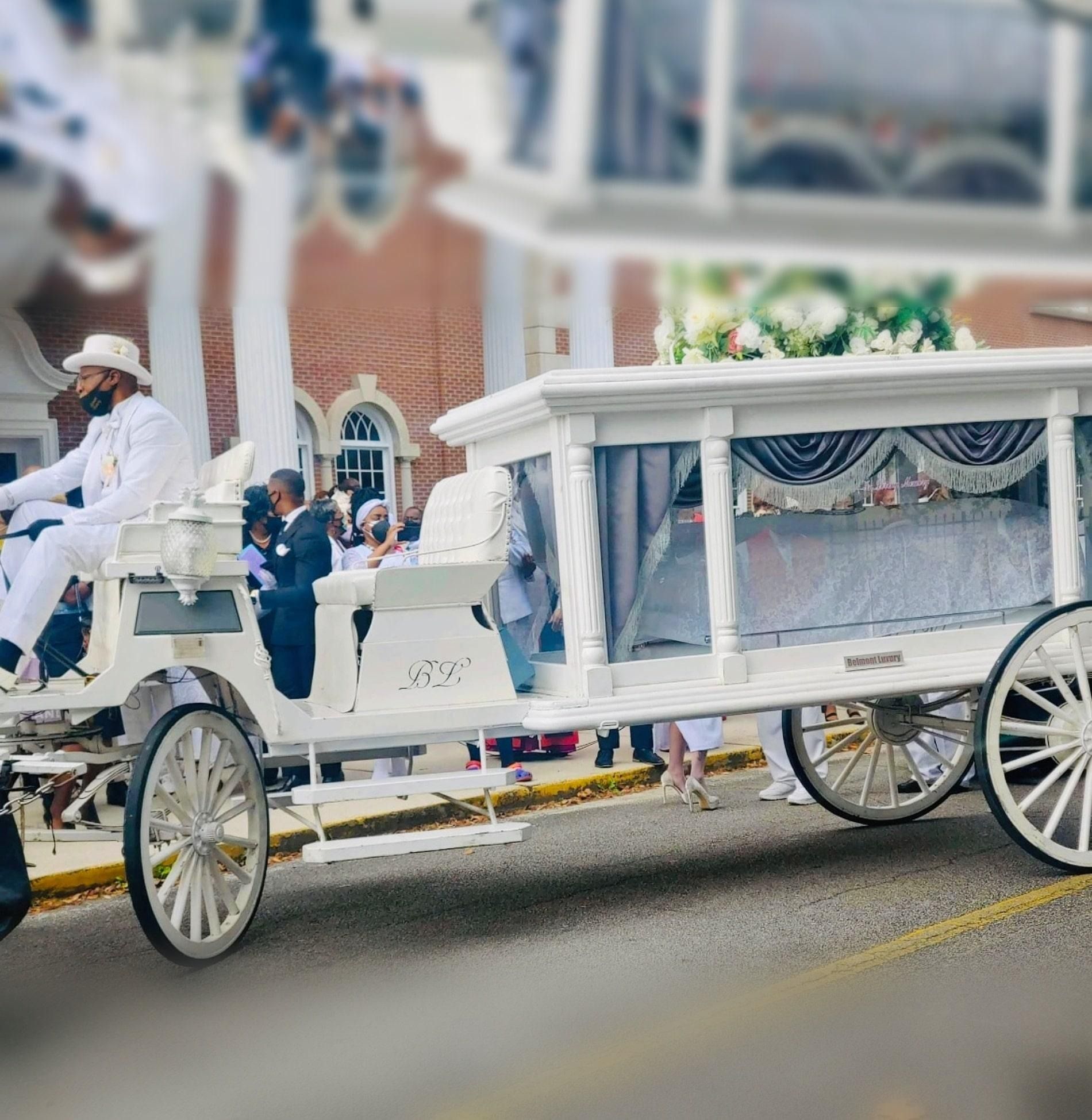 White horse-drawn carriage with draped canopy and white-clad passengers on a street, in front of a church.