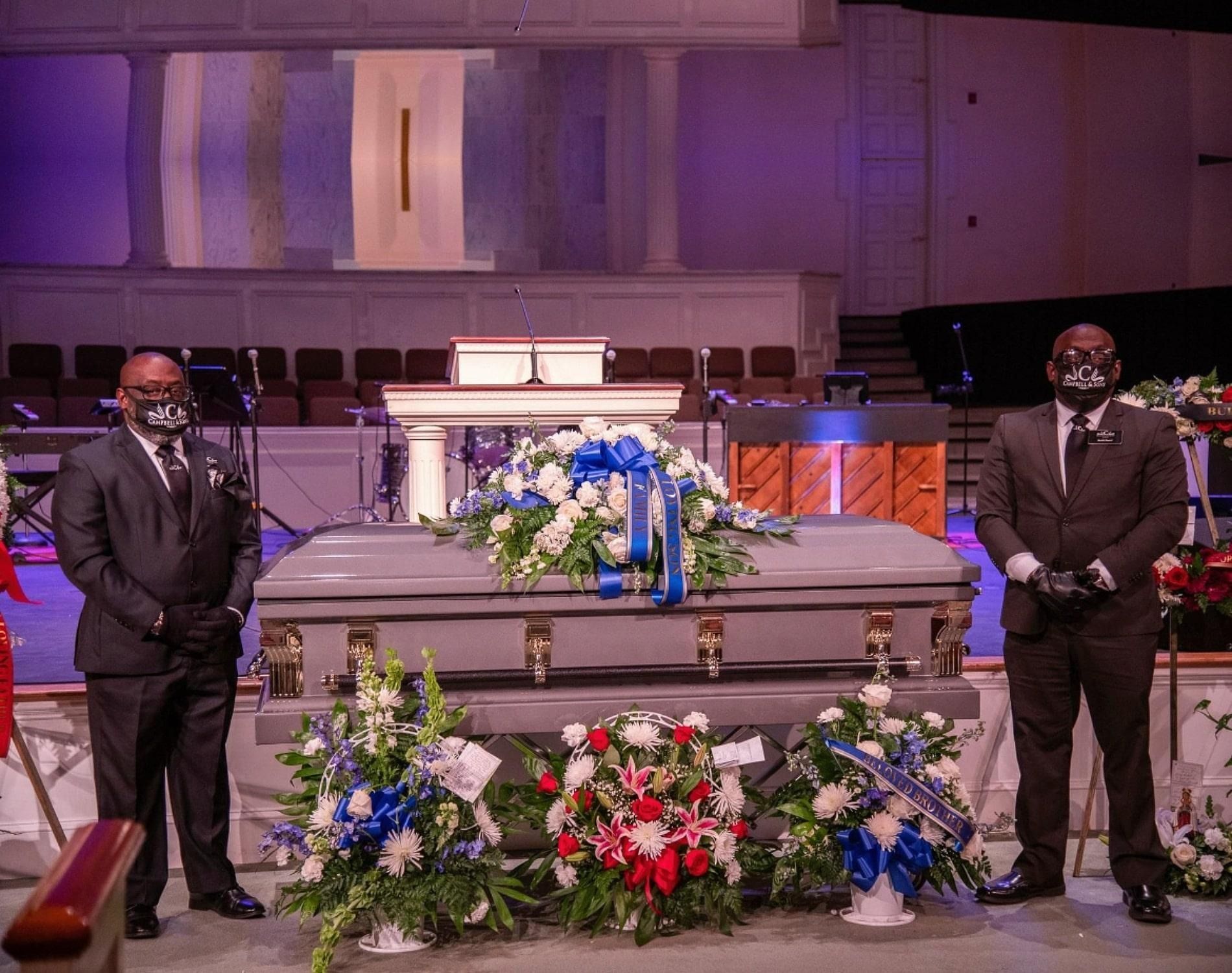 Two suited men stand beside a flower-covered casket in a church sanctuary.