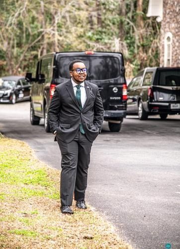 Man in a suit standing on a paved road beside parked vans, outdoors on a cloudy day