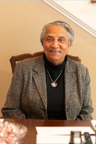 Smiling woman in a gray blazer seated indoors against a beige wall and staircase.