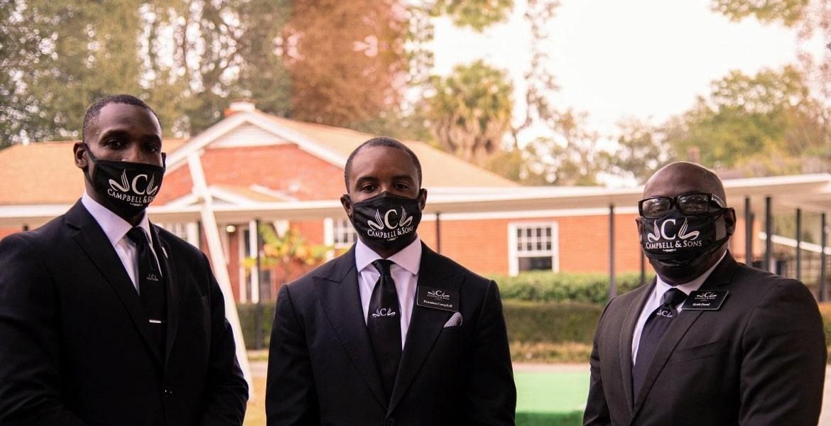 Three men in black suits and masks standing outdoors in front of a building.