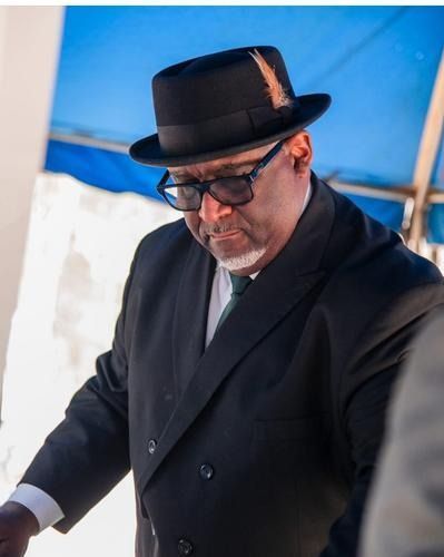 Man in a black suit and hat looking down, standing near a blue tent.