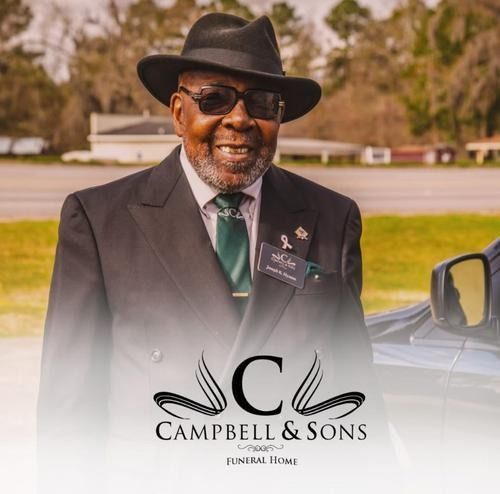 Man in black hat and suit standing outdoors beside a car, with Campbell & Sons Funeral Home logo.