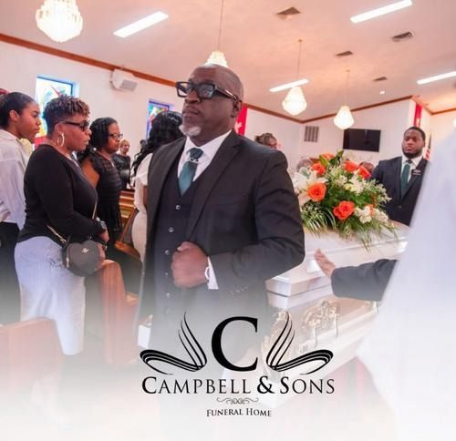 Funeral service with a suited man standing near flower arrangements and attendees in a bright chapel.