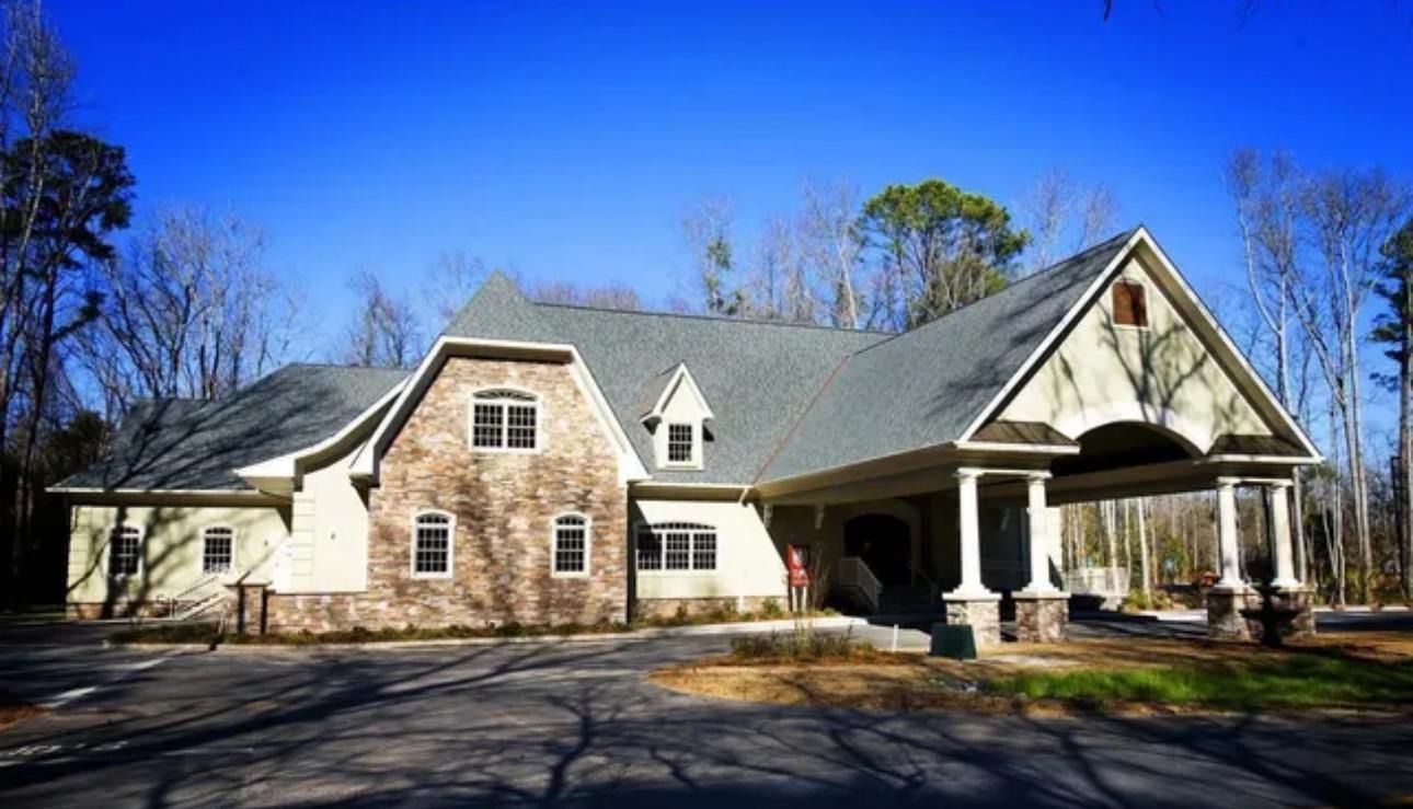 Stone house with a large covered porch, gabled roof, and bare trees under a bright blue sky