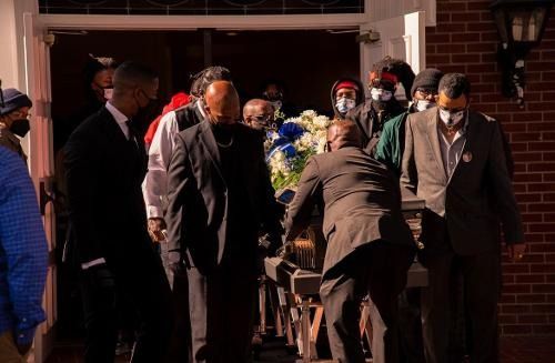 Mourners carry a casket draped with flowers through a church doorway, surrounded by suit-clad attendants.