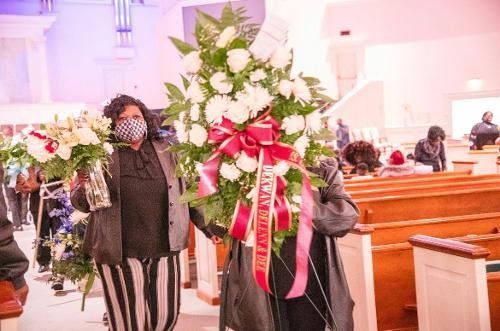 People carrying large white floral arrangements with pink ribbons inside a church