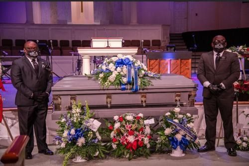 Two men in suits stand beside a flower-covered casket in a church sanctuary.
