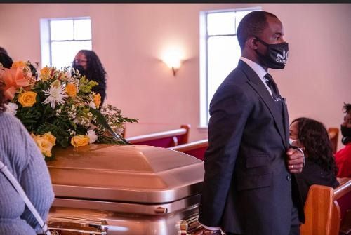 Funeral service with a gold casket and floral arrangement, mourners seated nearby