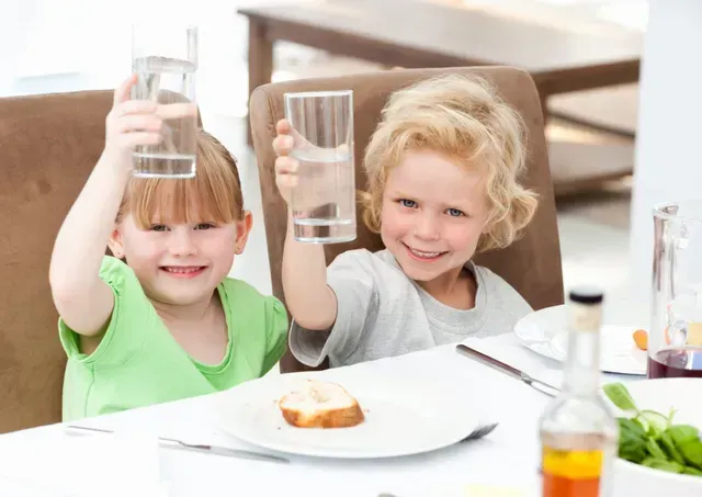 A boy and a girl are sitting at a table holding up glasses of water.
