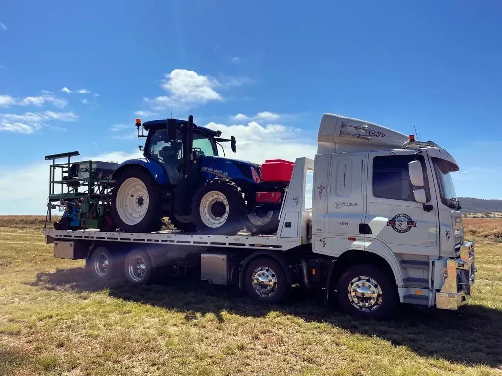 A White Flatbed Tow Truck Transporting a Blue Tractor — Tamworth Tilt Trays in Tamworth, NSW