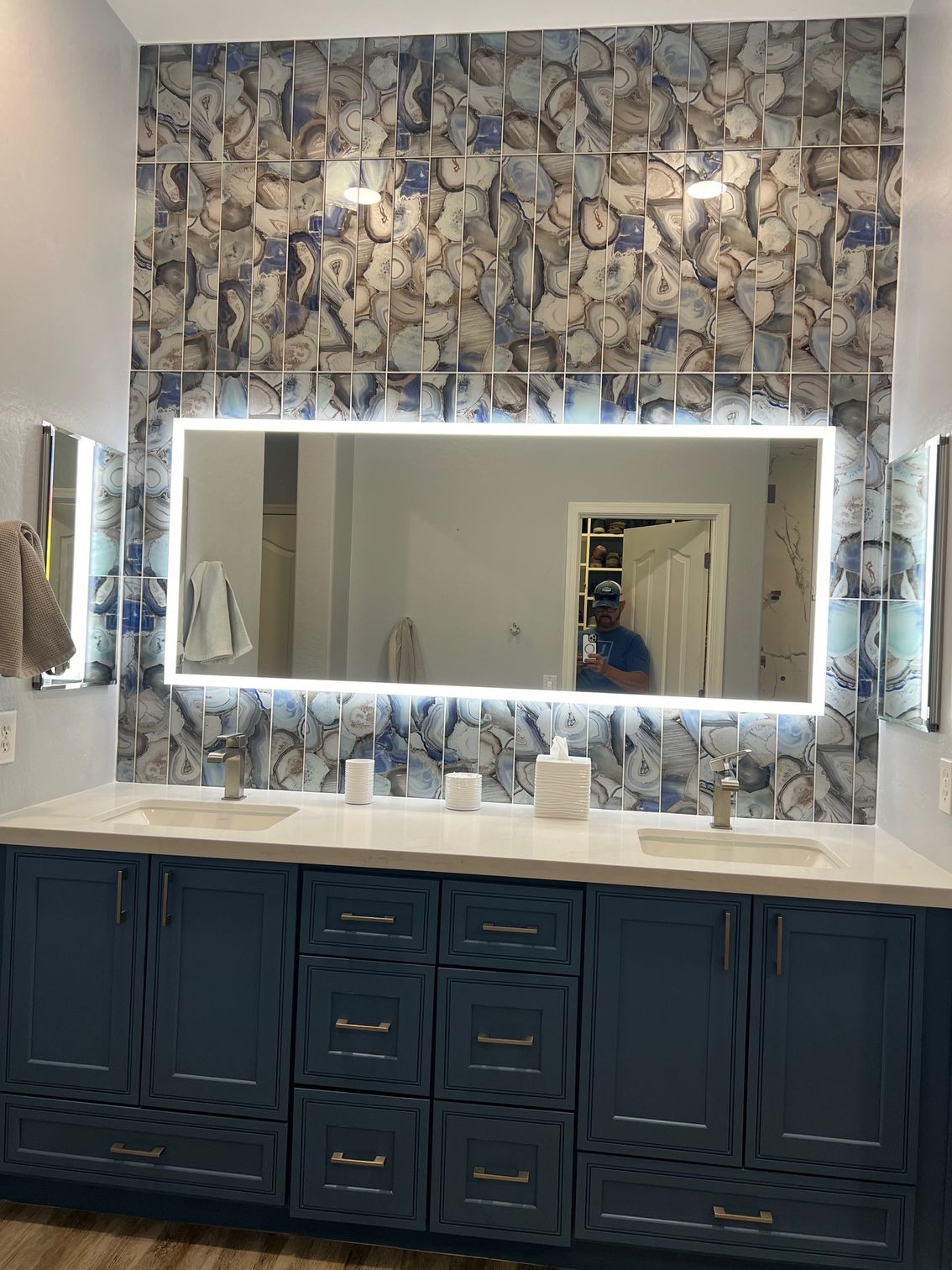 Bathroom vanity with large backlit mirror, glass feature wall, and dark blue refinished cabinets