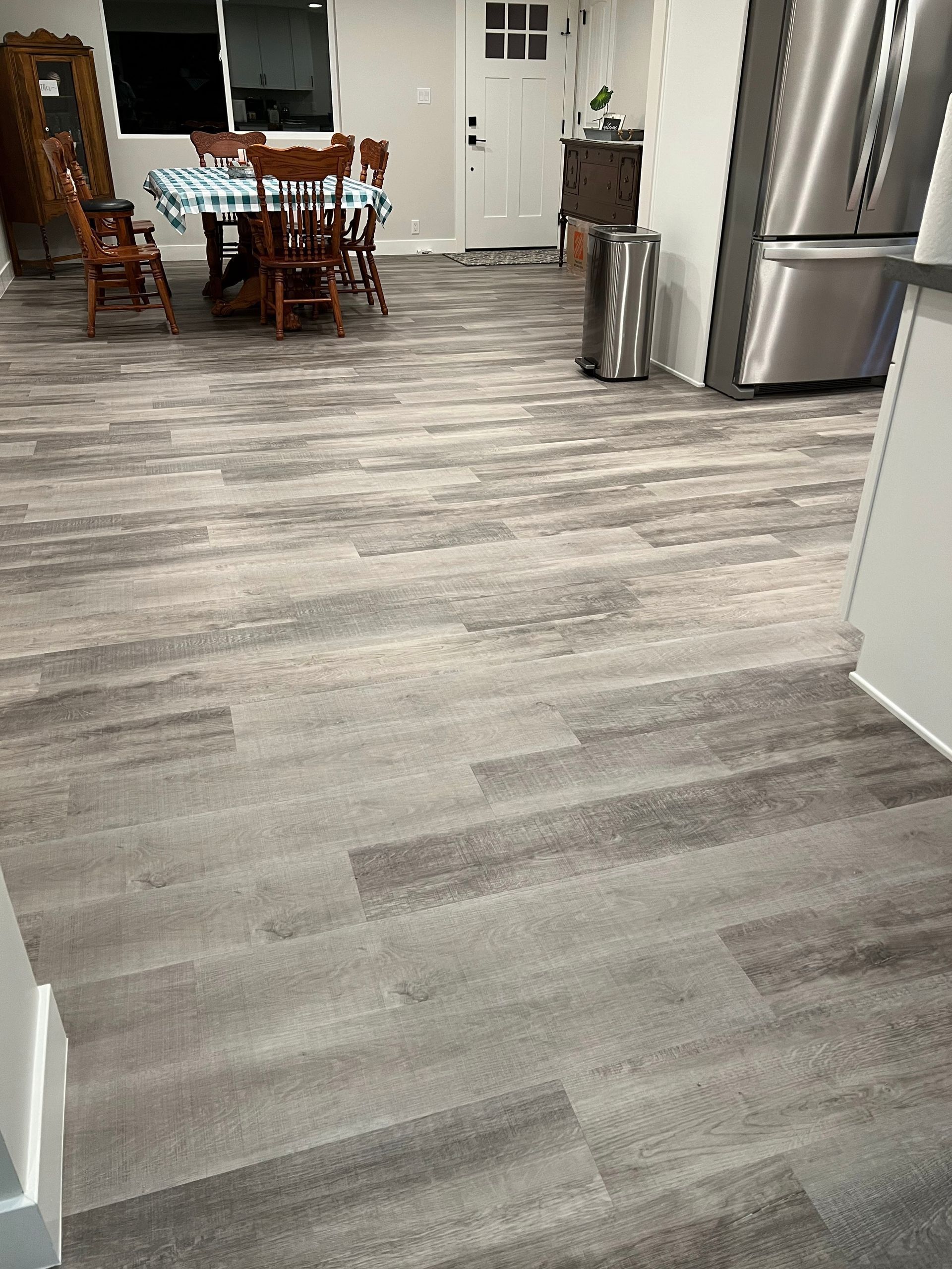 Modern kitchen with gray wood-look flooring, dining table, white cabinets, and stainless steel refrigerator.