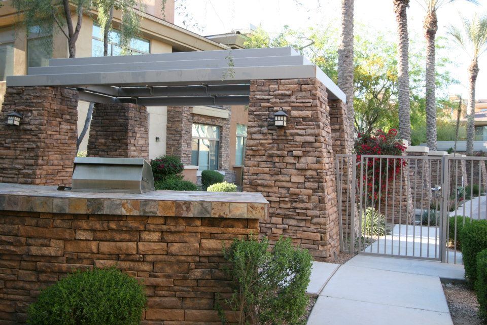 Stone entry wall with a service window and metal gate beside a sidewalk in a landscaped courtyard