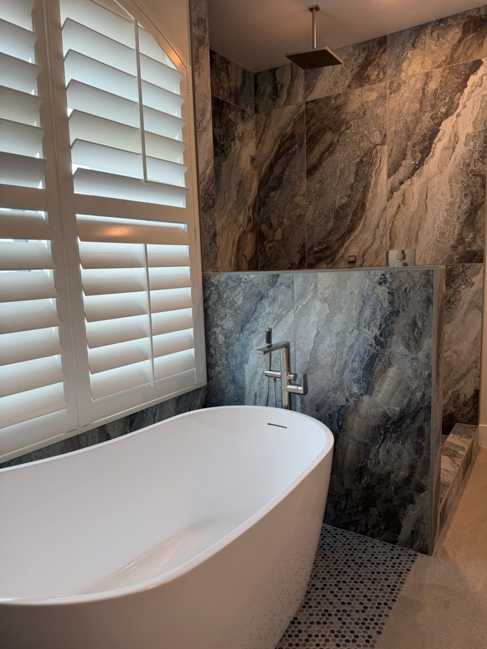 Modern bathroom with freestanding tub, gray stone shower wall, and white plantation shutters
