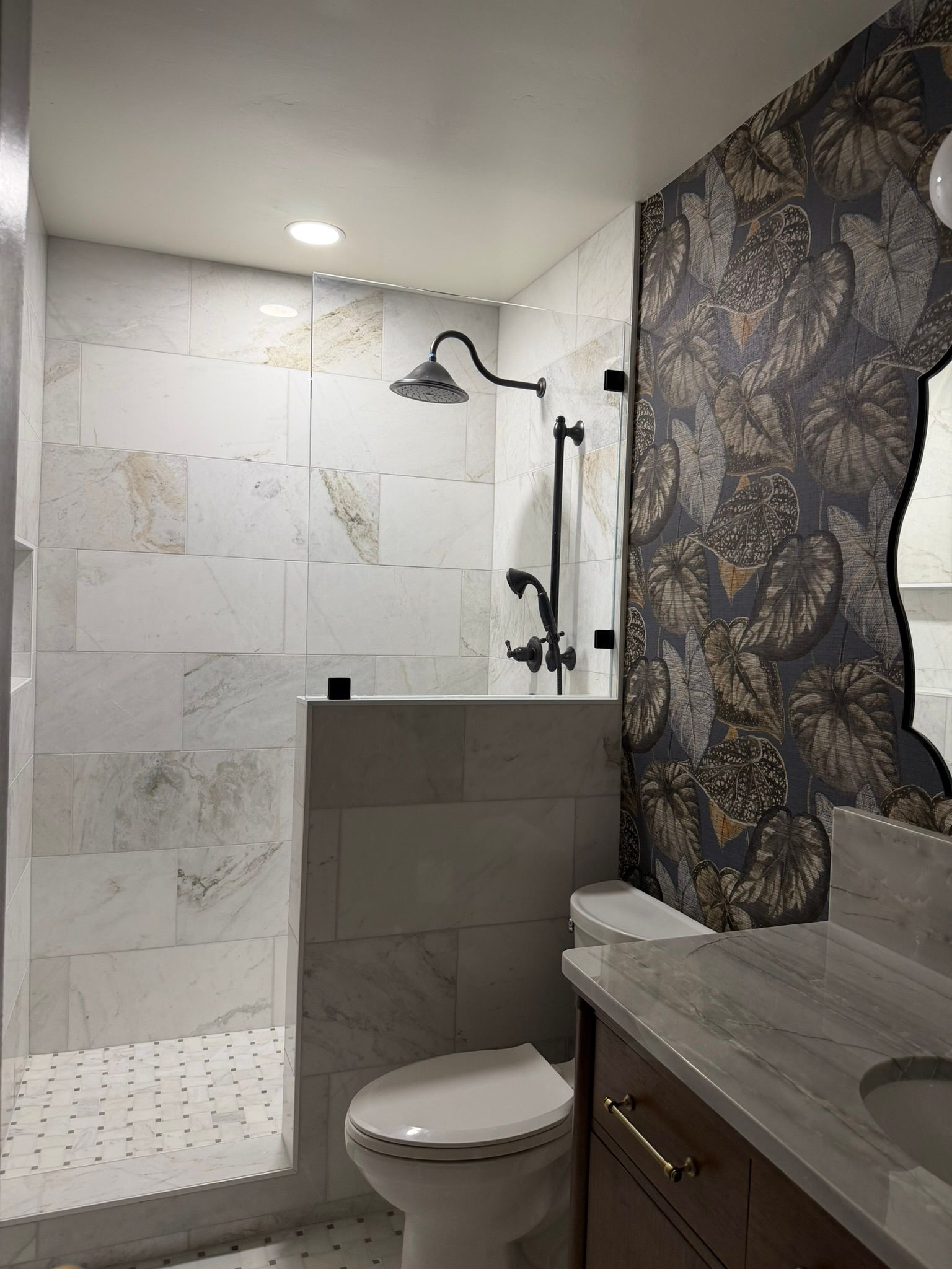 Modern bathroom with tiled shower, rainfall showerhead, toilet, sink, and dark floral wallpaper accent wall