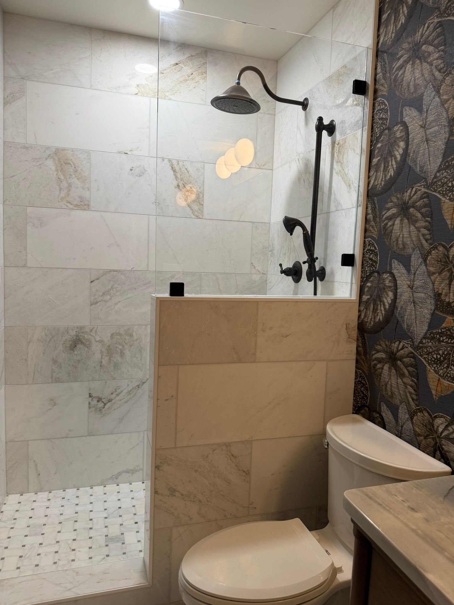 Shower stall with tiled walls, glass door, and a toilet beside a beige half wall.
