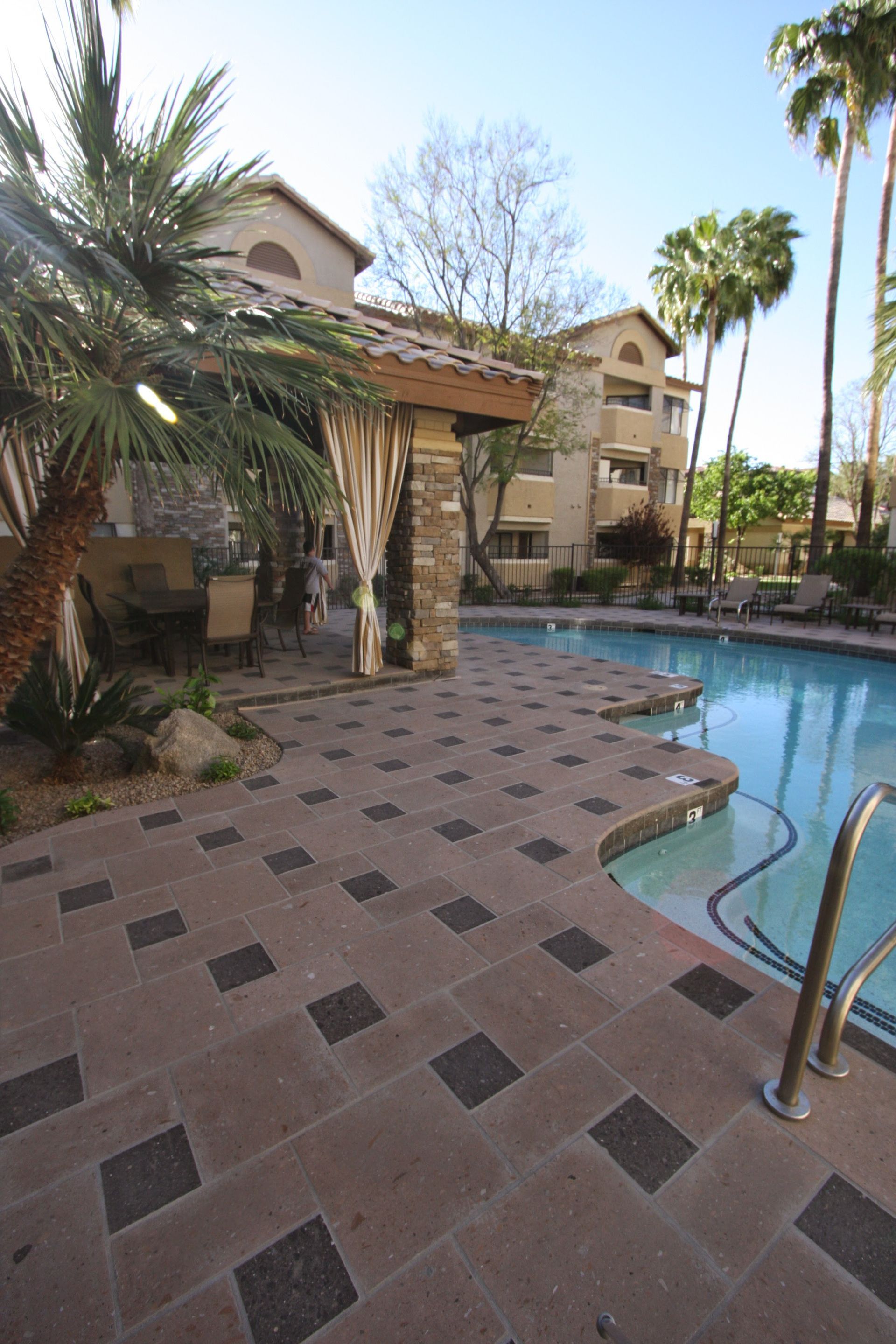 Apartment pool area with curved blue pool, palm trees, and tan tiled patio.