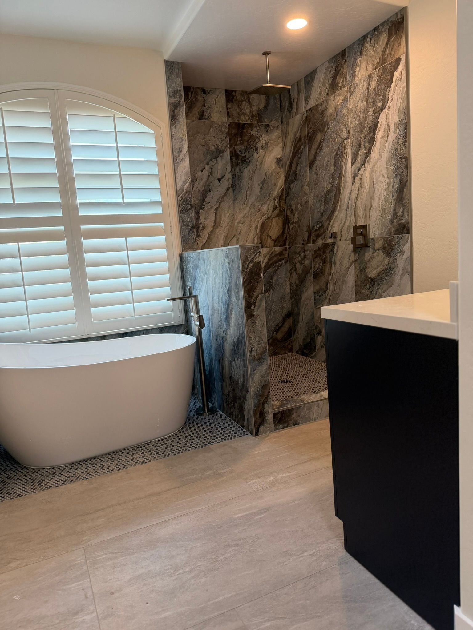 Modern bathroom with freestanding tub, marble shower wall, white shutters, and black vanity