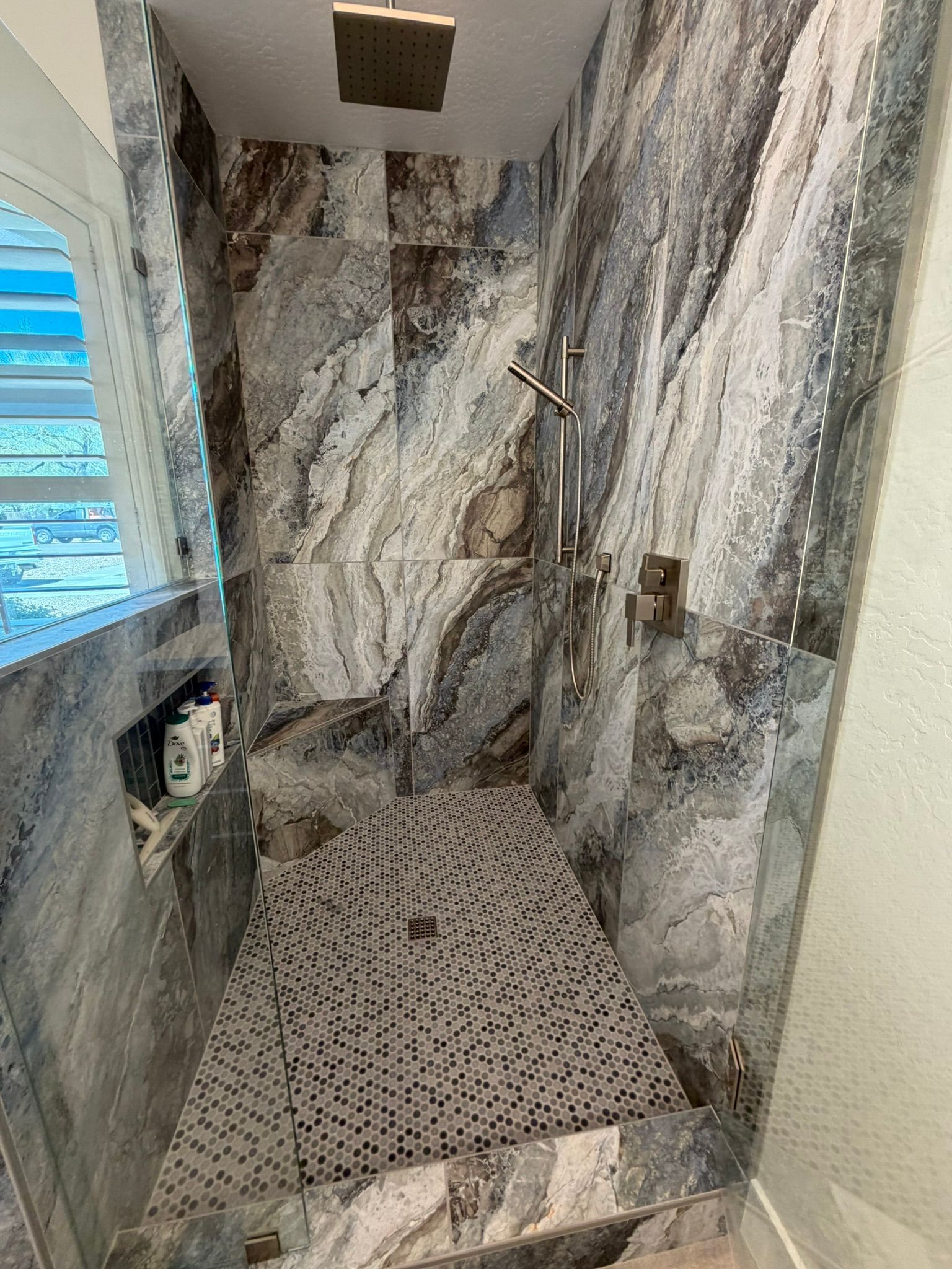 Marble-tiled shower with wall-mounted fixtures, window, and pebble-pattern floor