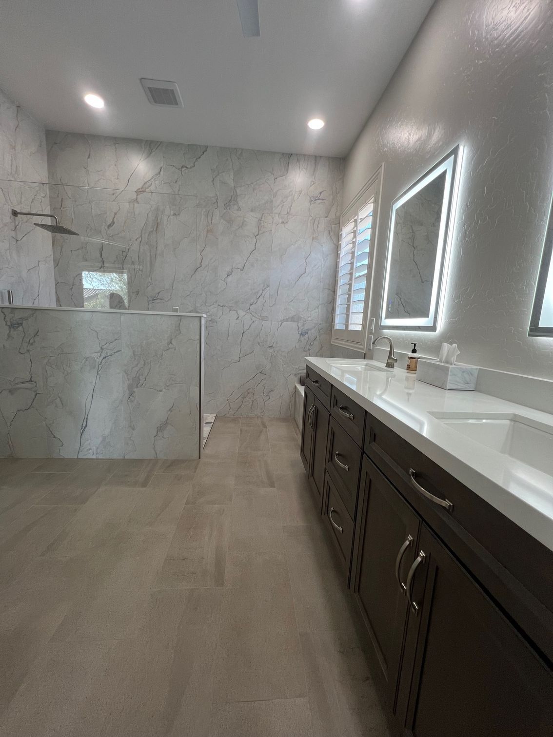Modern marble bathroom with dark vanity, double sinks, large mirror, and glass shower.
