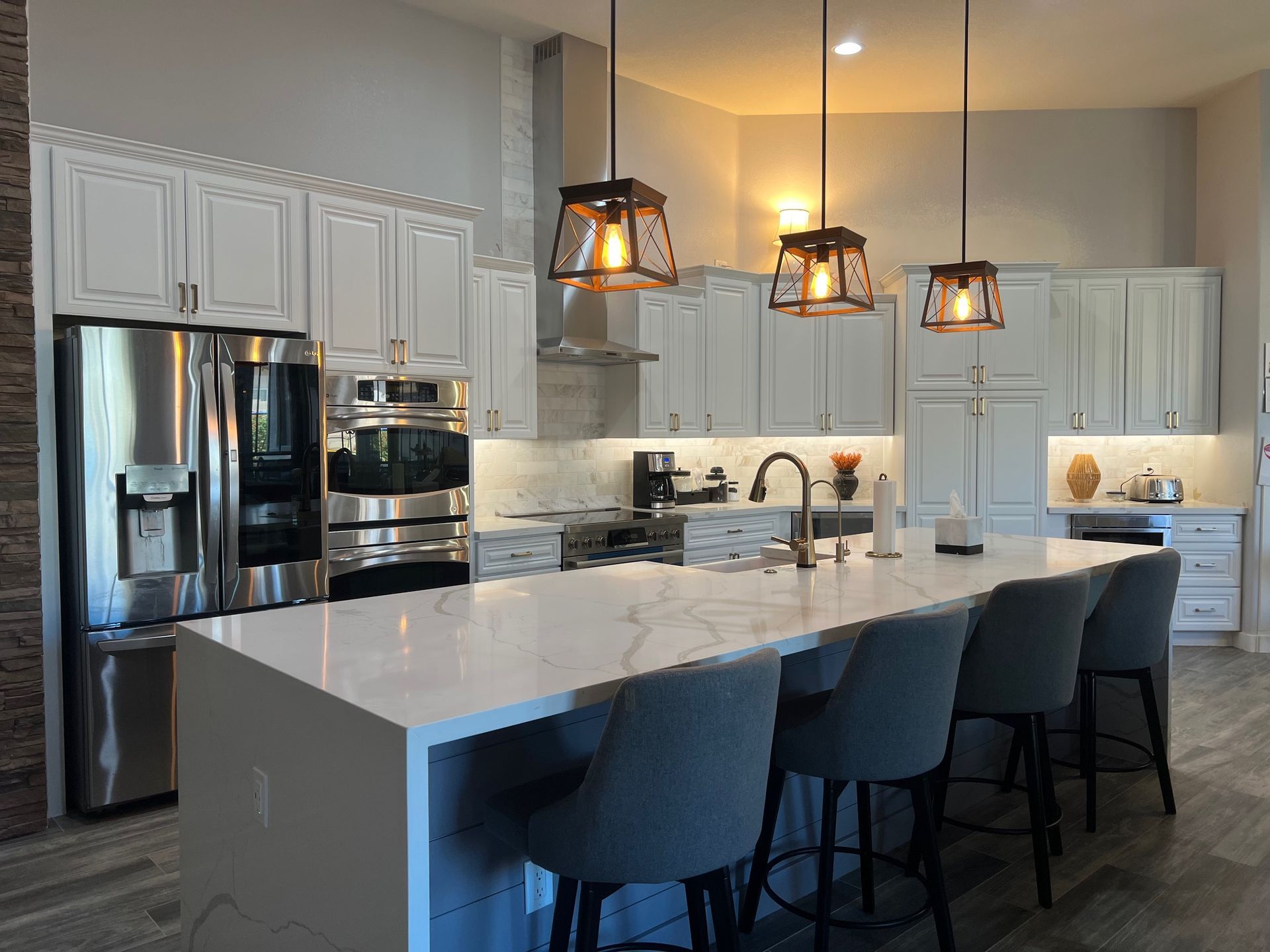 Modern white kitchen with large island, stainless steel appliances, pendant lights, and four dark barstools