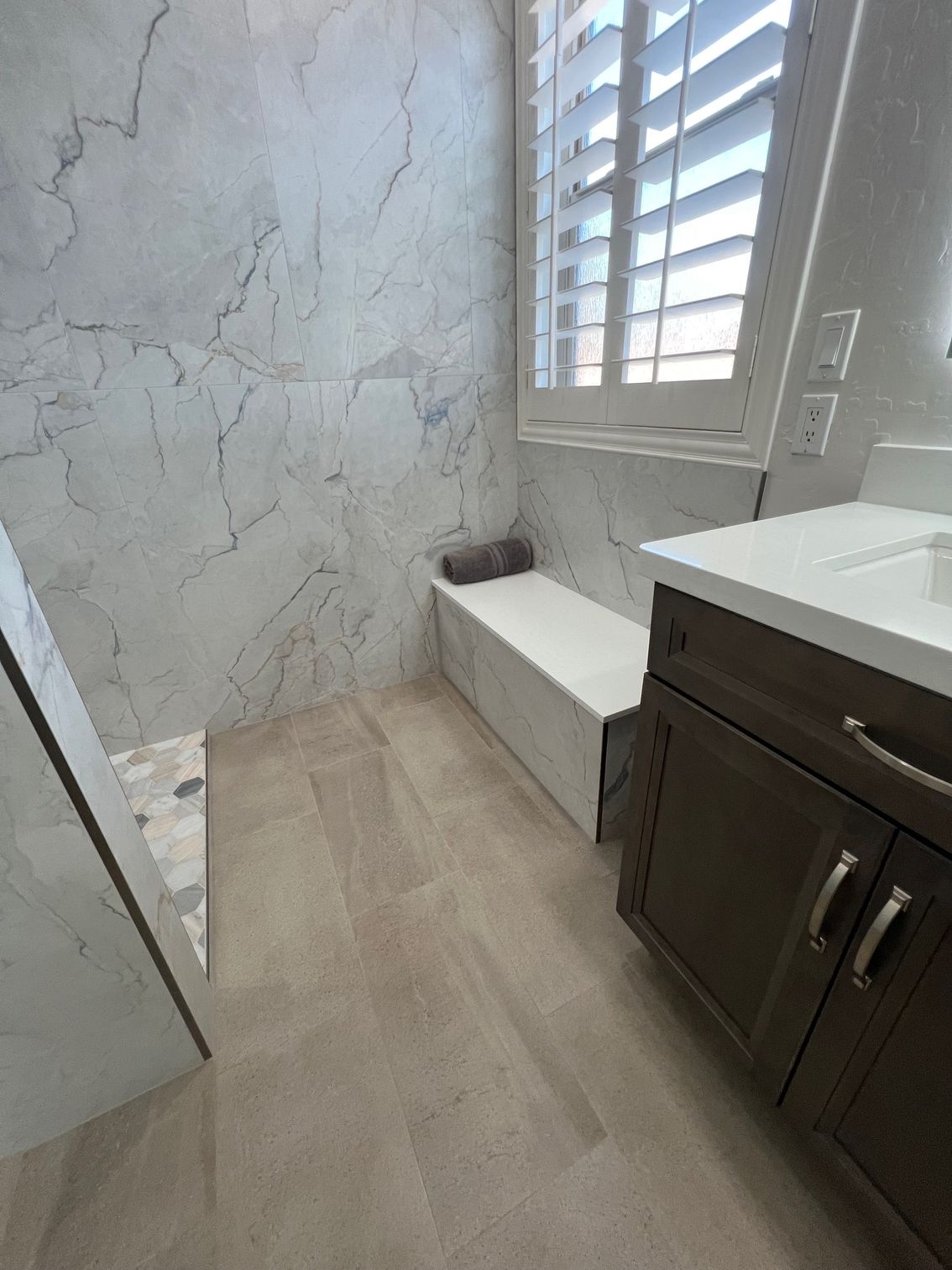 Modern bathroom with marble-look wall, white bench, dark vanity, and window shutters