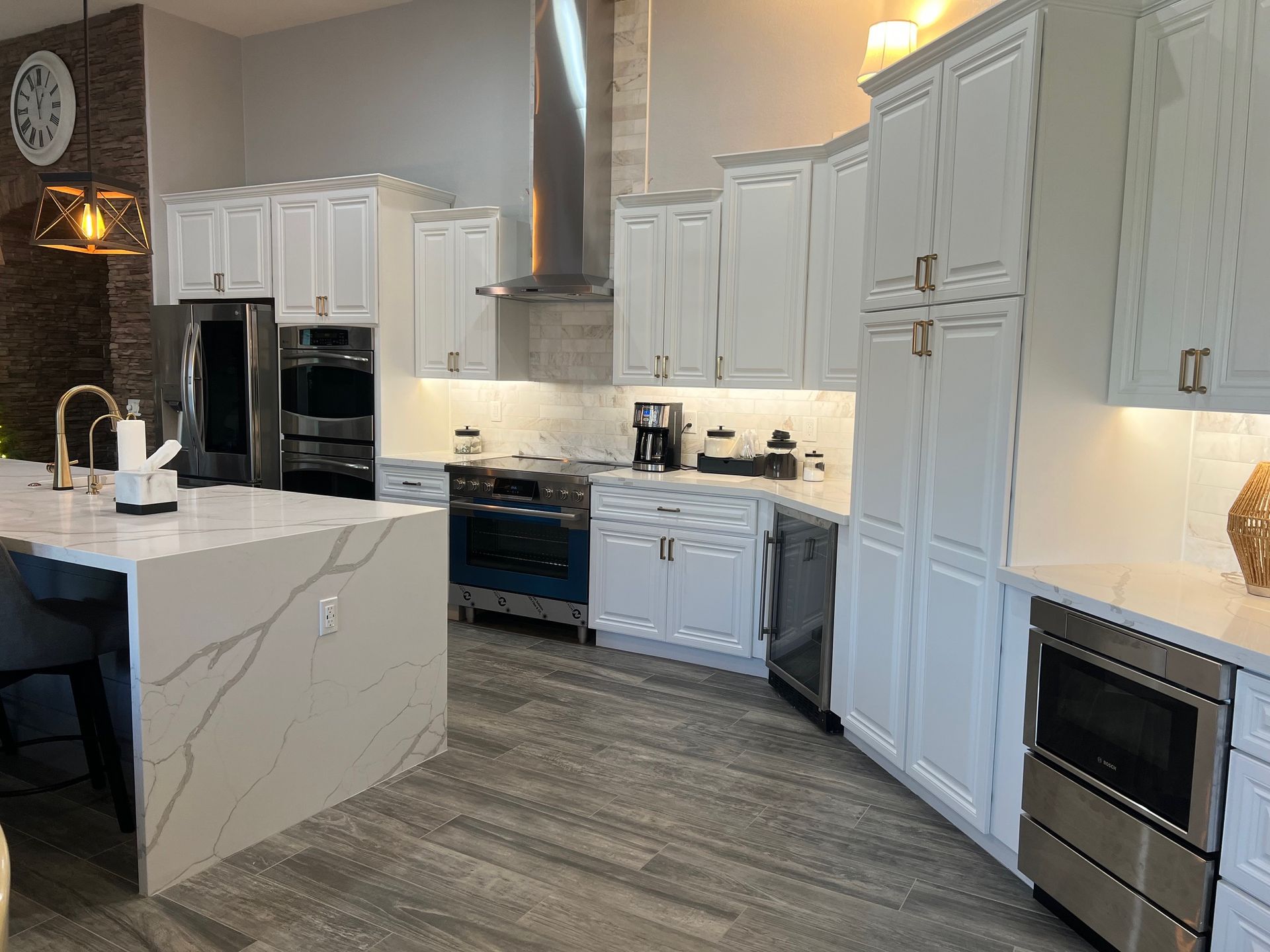 Modern white kitchen with marble island, stainless appliances, gray wood floor, and pendant lighting