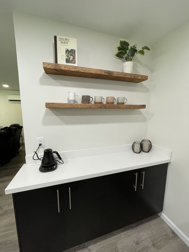 Minimal kitchen coffee station with black cabinets, white countertop, floating shelves, kettle, mugs, and plant.