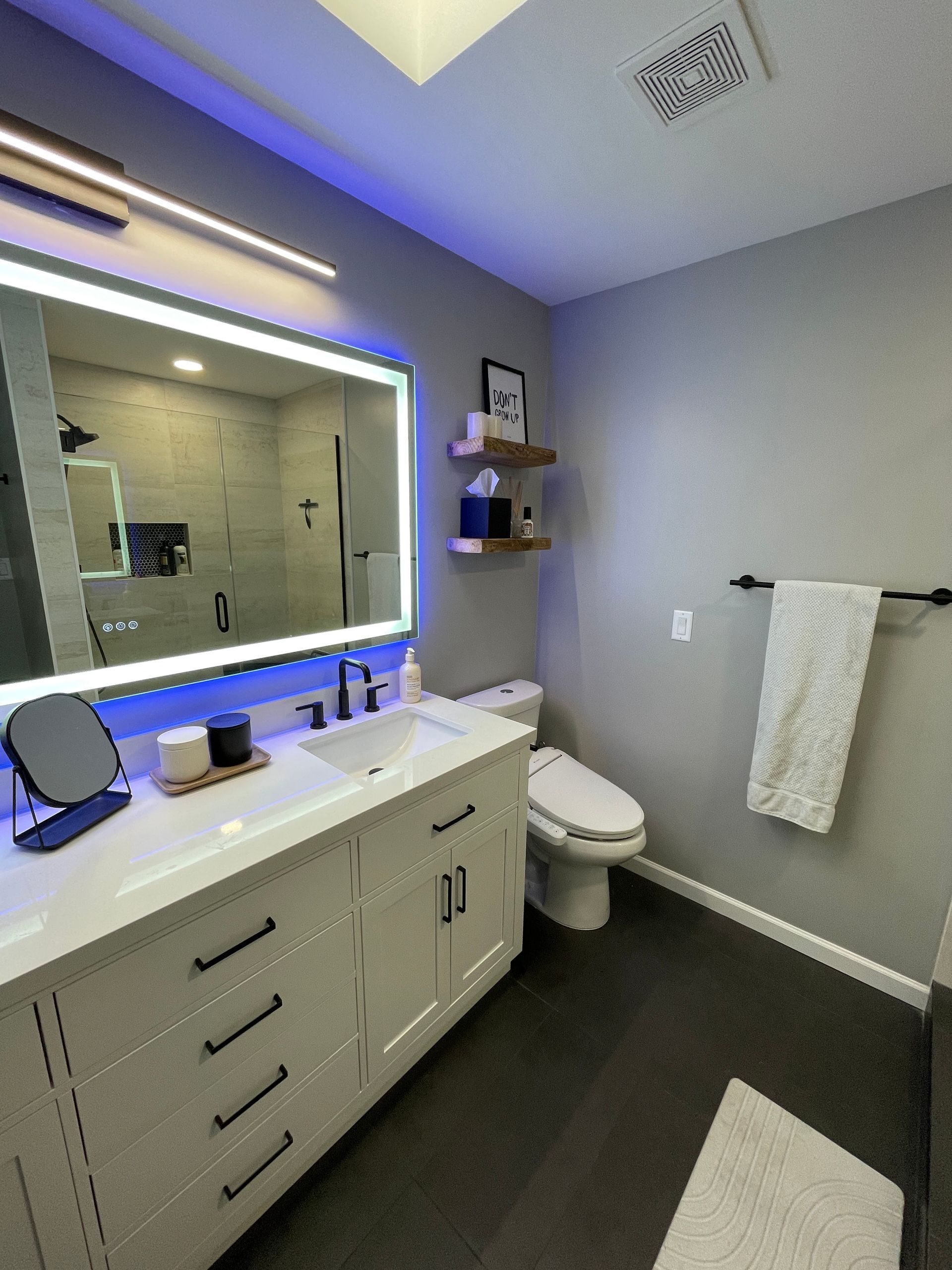 Modern bathroom with illuminated mirror, white vanity, toilet, towel rack, and dark floor.