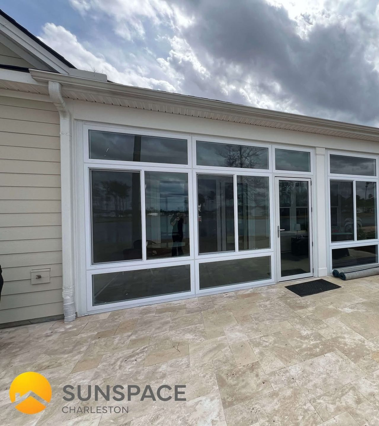 Sunroom with large glass windows and white frames on a beige patio. The sky is cloudy.