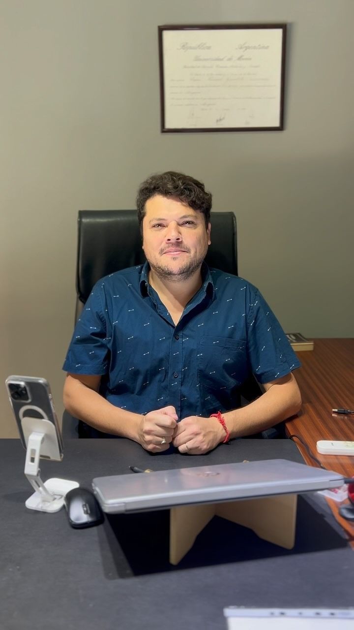 Hombre con camisa azul sentado en un escritorio con un ordenador portátil, ratón, teléfono y diploma al fondo, sonriendo.