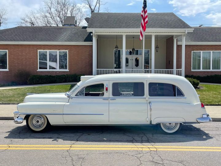 White vintage hearse parked on a street in front of a brick building with an American flag.