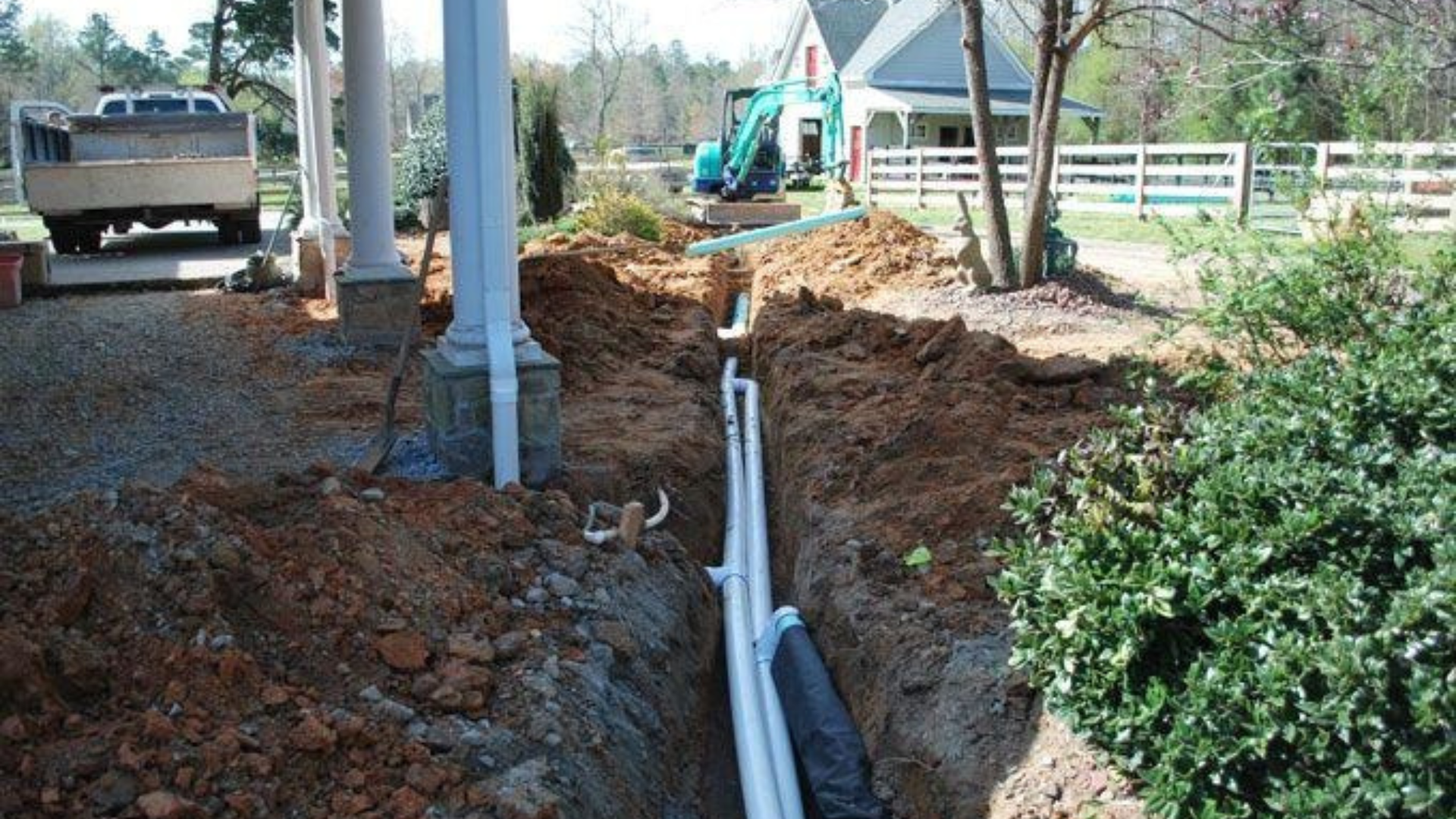 Trenching work with exposed utility pipes running through a muddy yard beside a house and truck