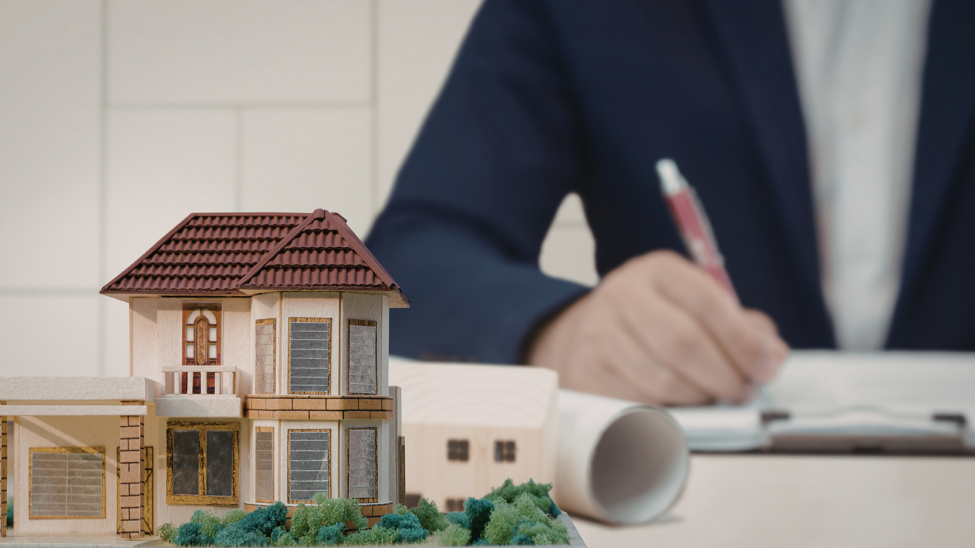 Scale model house with rolled blueprint in foreground and person writing in blurred background