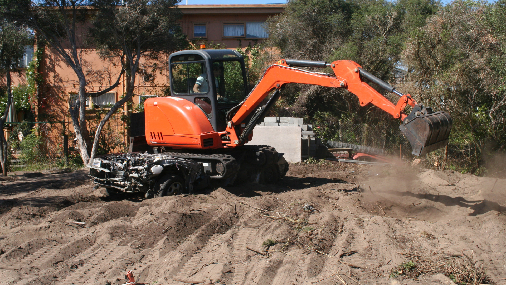 Orange excavator digging dirt in a yard near a house, with soil piled around it.