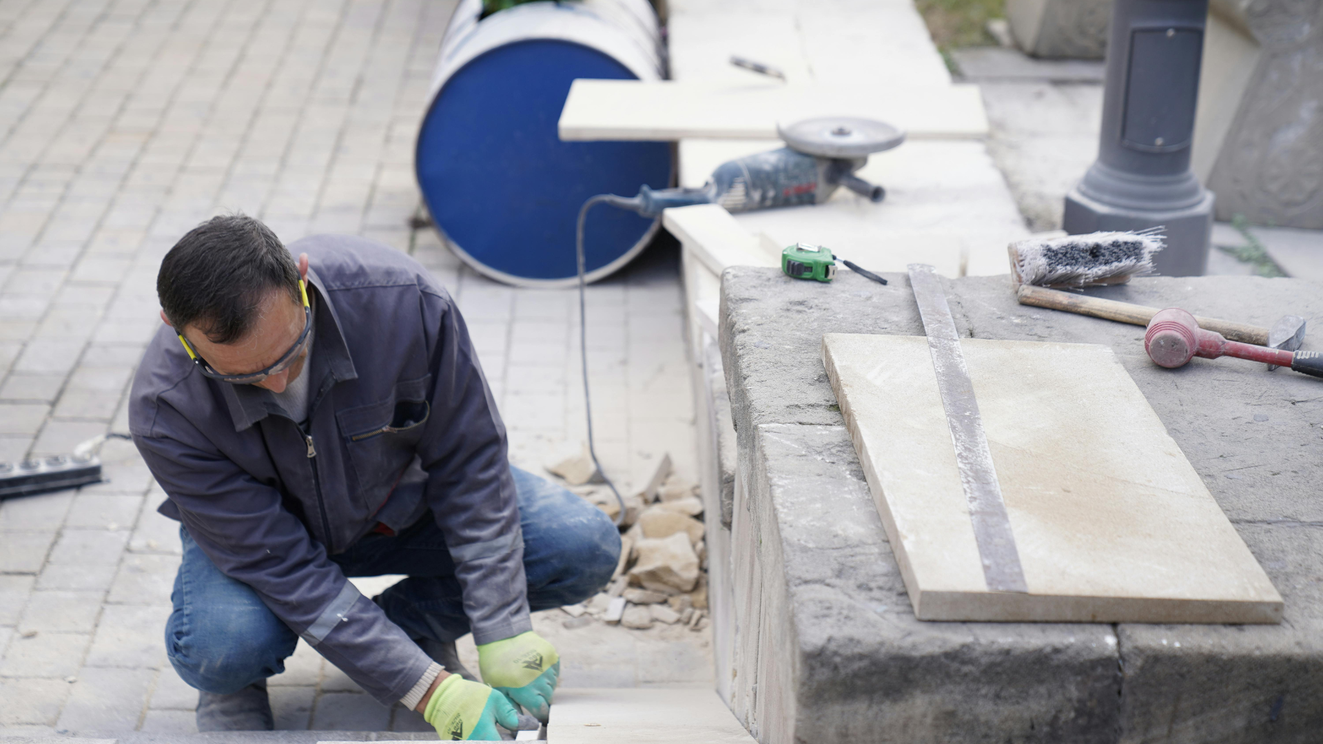 Worker in gloves kneels outdoors, cutting stone slabs beside a concrete block and construction tools.